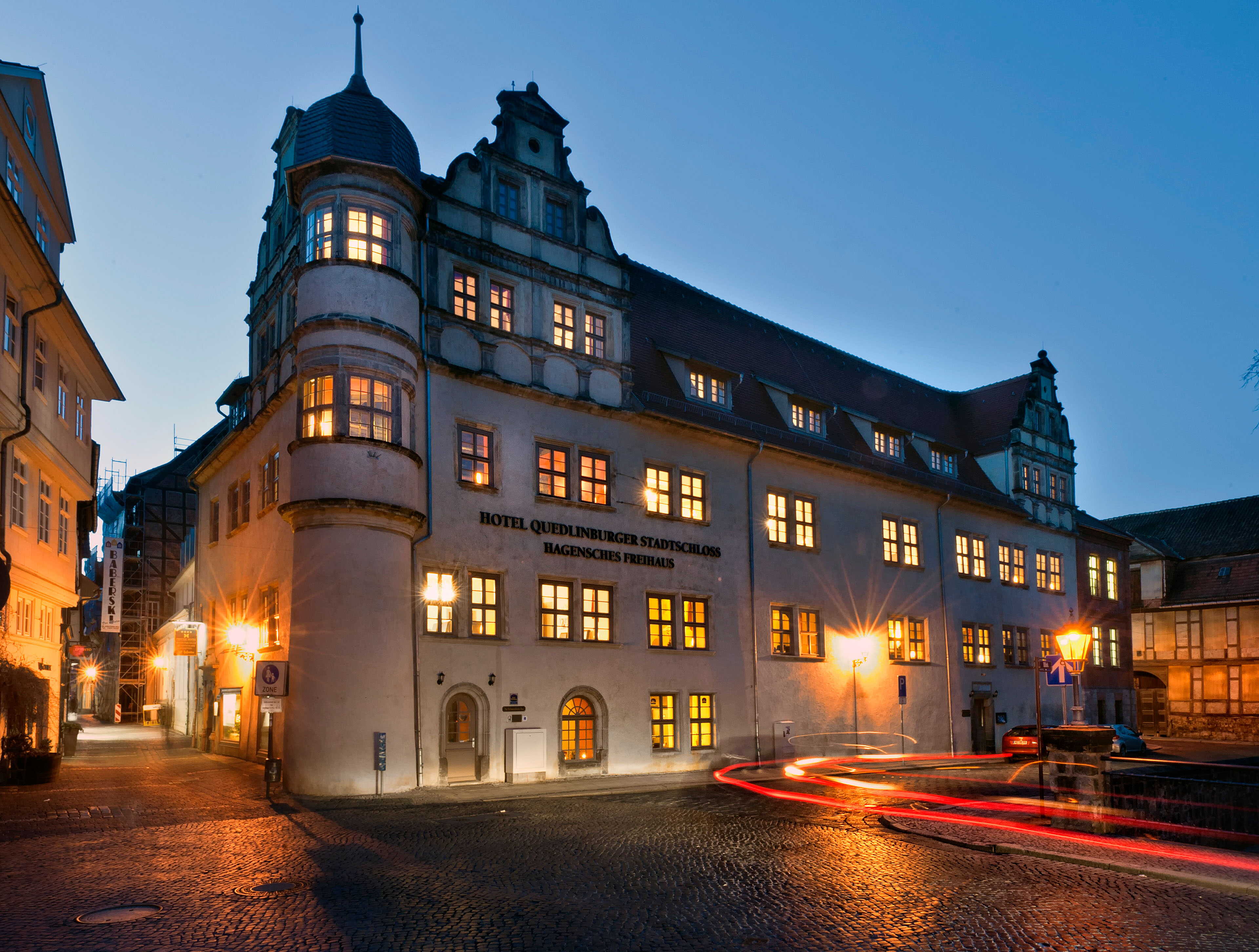 The illuminated exterior of Hotel Quedlinburger Stadtschloss glows warmly at dusk with blurred car lights on the cobblestone street.