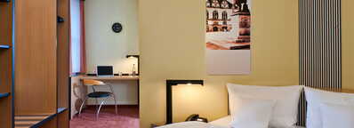 Ein modernes Business-Zimmer mit Schreibtisch, Laptop und gemütlichem Bett wartet in einem hellen Raum auf Gäste.