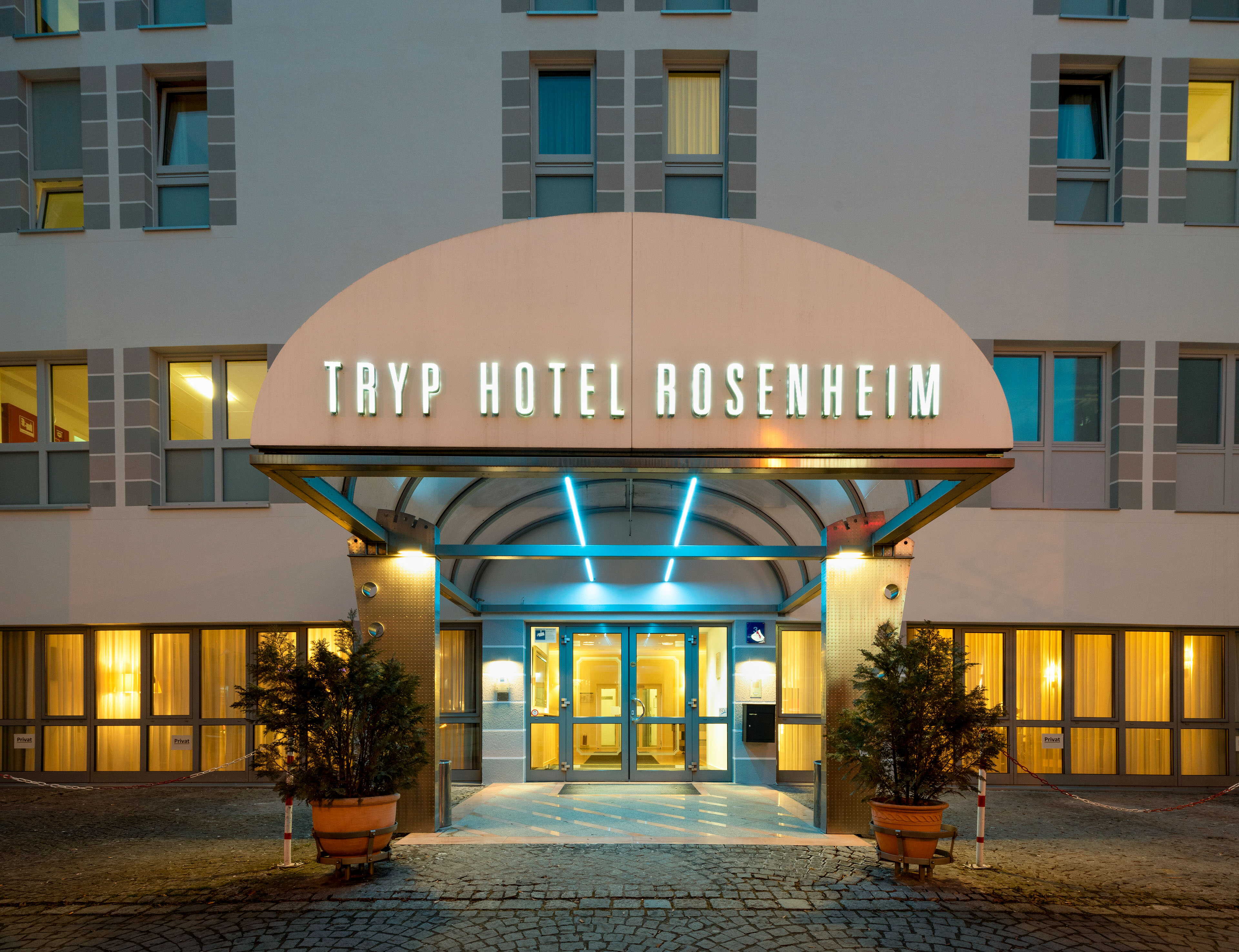 The illuminated entrance of Tryp Hotel Rosenheim glows warmly at dusk, flanked by potted plants and large windows.