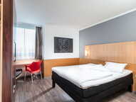 A modern single room features a cozy bed, a bright red chair by the window, and a stylish wall art piece.