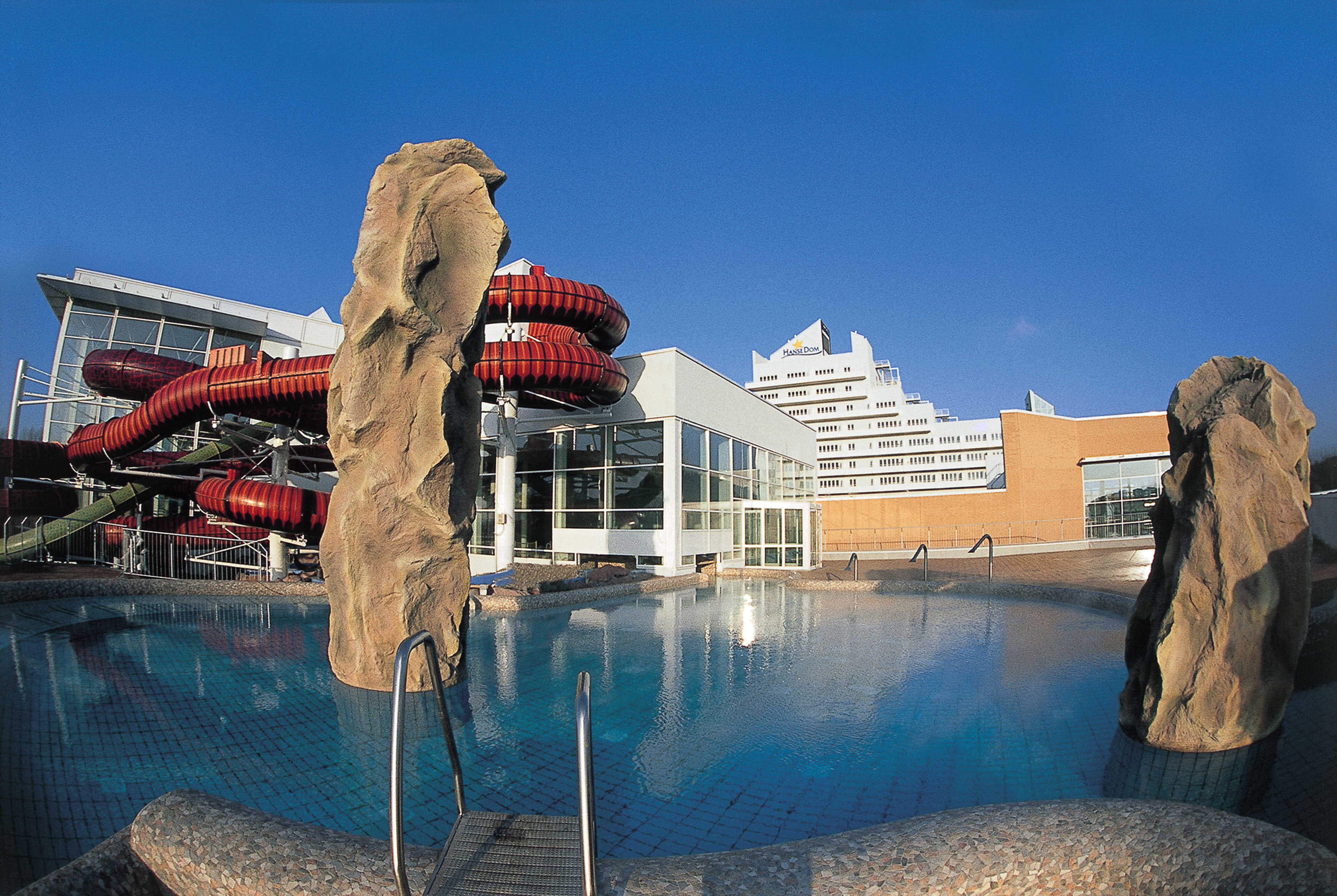 A modern water park features winding red slides, rocky sculptures, and a large pool under a clear blue sky.