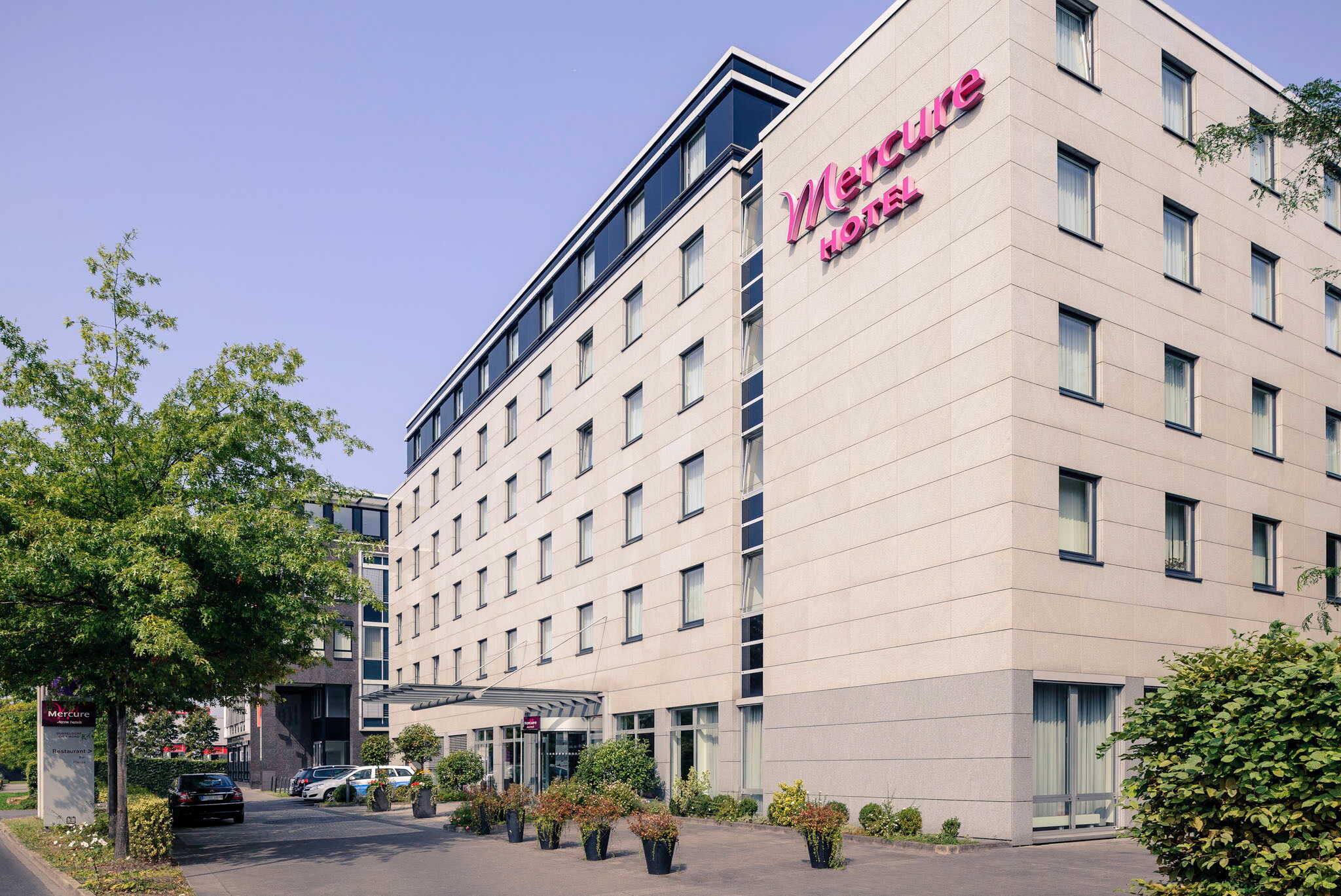 The Mercure Hotel building stands prominently under a clear sky, surrounded by trees and parked cars.