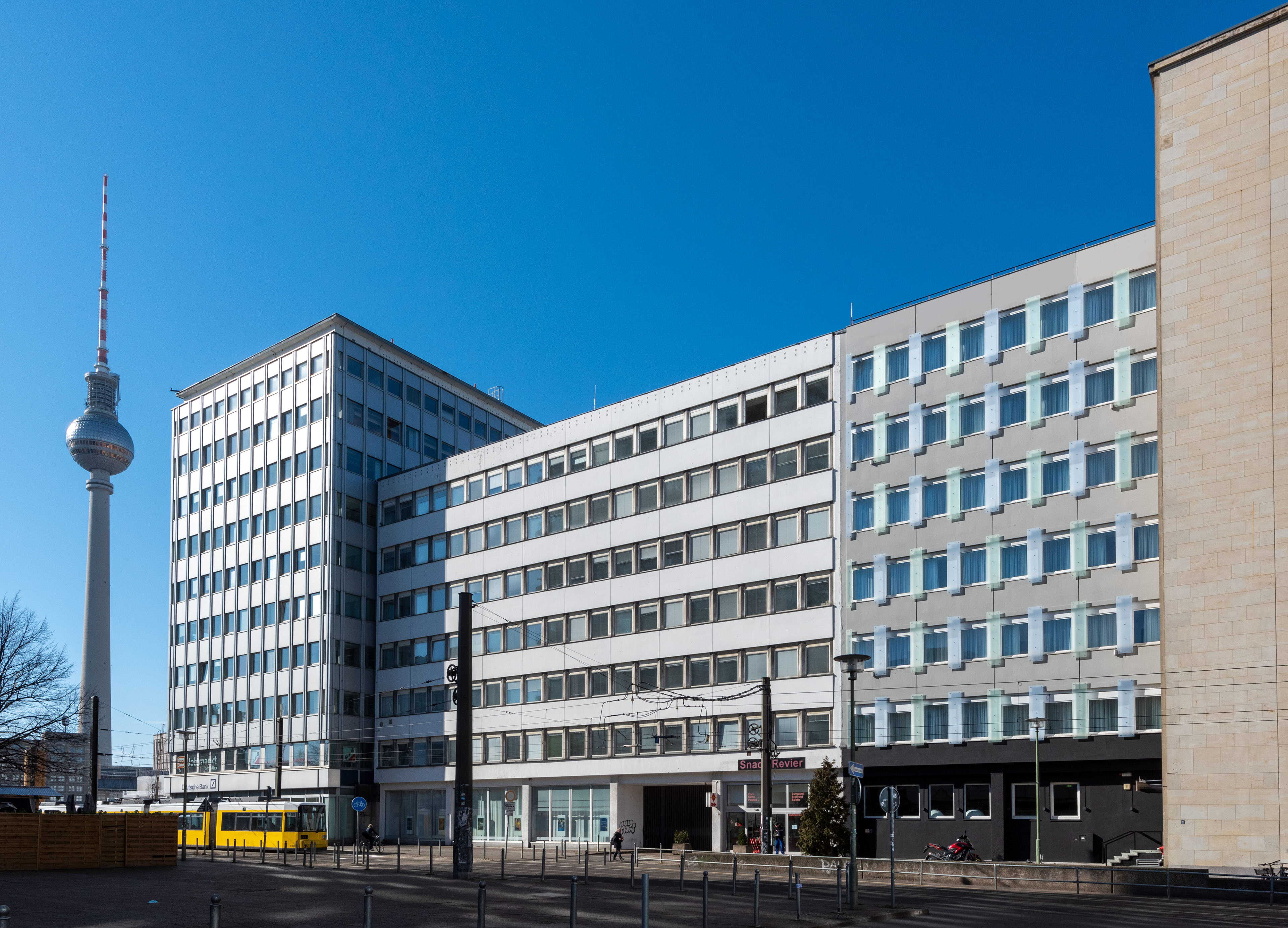 The exterior of a modern building in Berlin is shown against a clear blue sky, with the TV tower visible in the background.