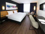 A modern hotel room features a sleek double bed, abstract art, and a workspace with a smooth desk and chair.