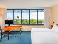 A bright hotel room features twin beds, a desk with a TV, and a large window offering a scenic view of trees and a distant cityscape.