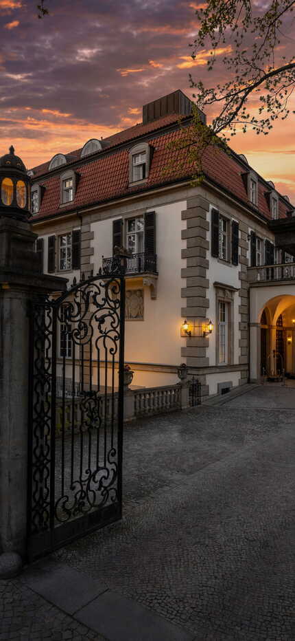 A grand hotel entrance is warmly illuminated by evening sunlight under a vibrant sunset sky.