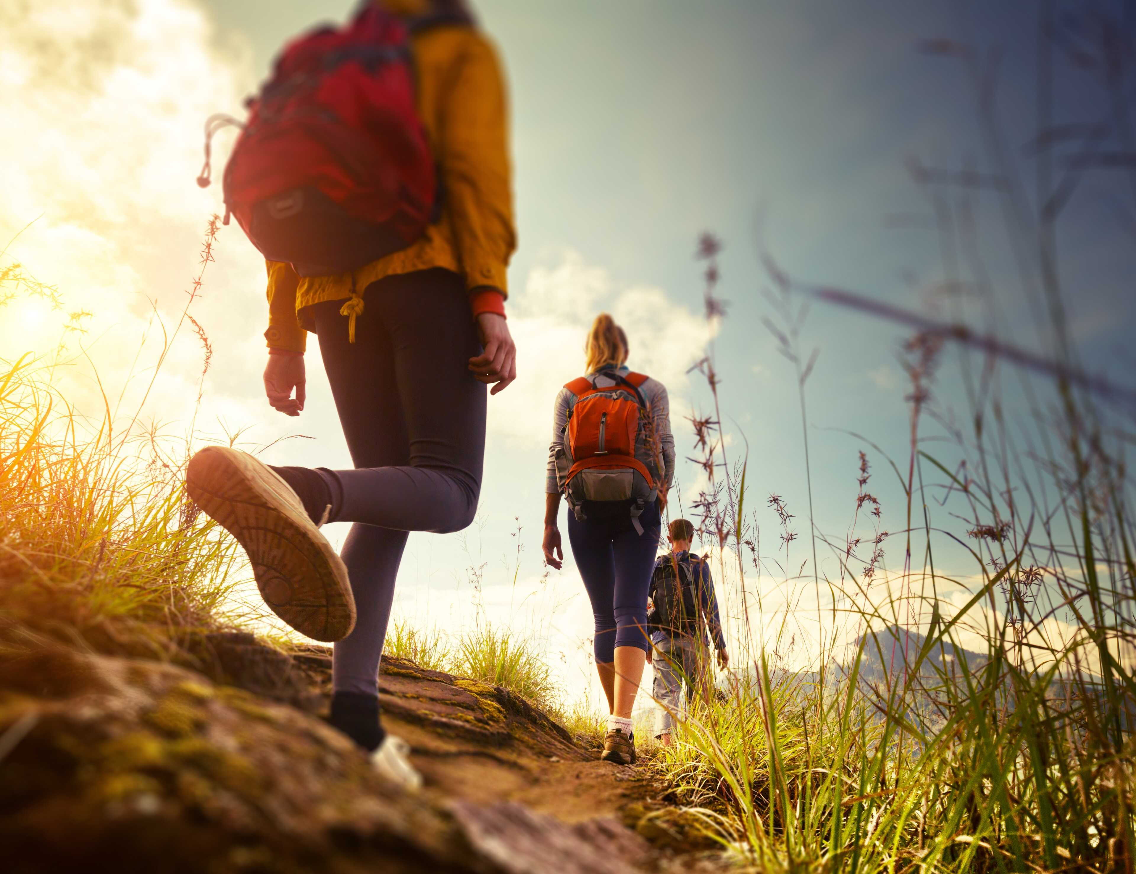 Drei Wanderer mit Rucksäcken gehen bei Sonnenuntergang einen grasbewachsenen Pfad entlang, die Natur in Peine erkundend.