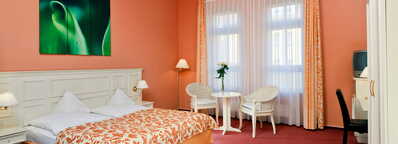 Gemütliches Zimmer mit einem großen Bett, einem kleinen Tisch mit zwei Stühlen und einem beruhigenden grünen Kunstwerk an der Wand.