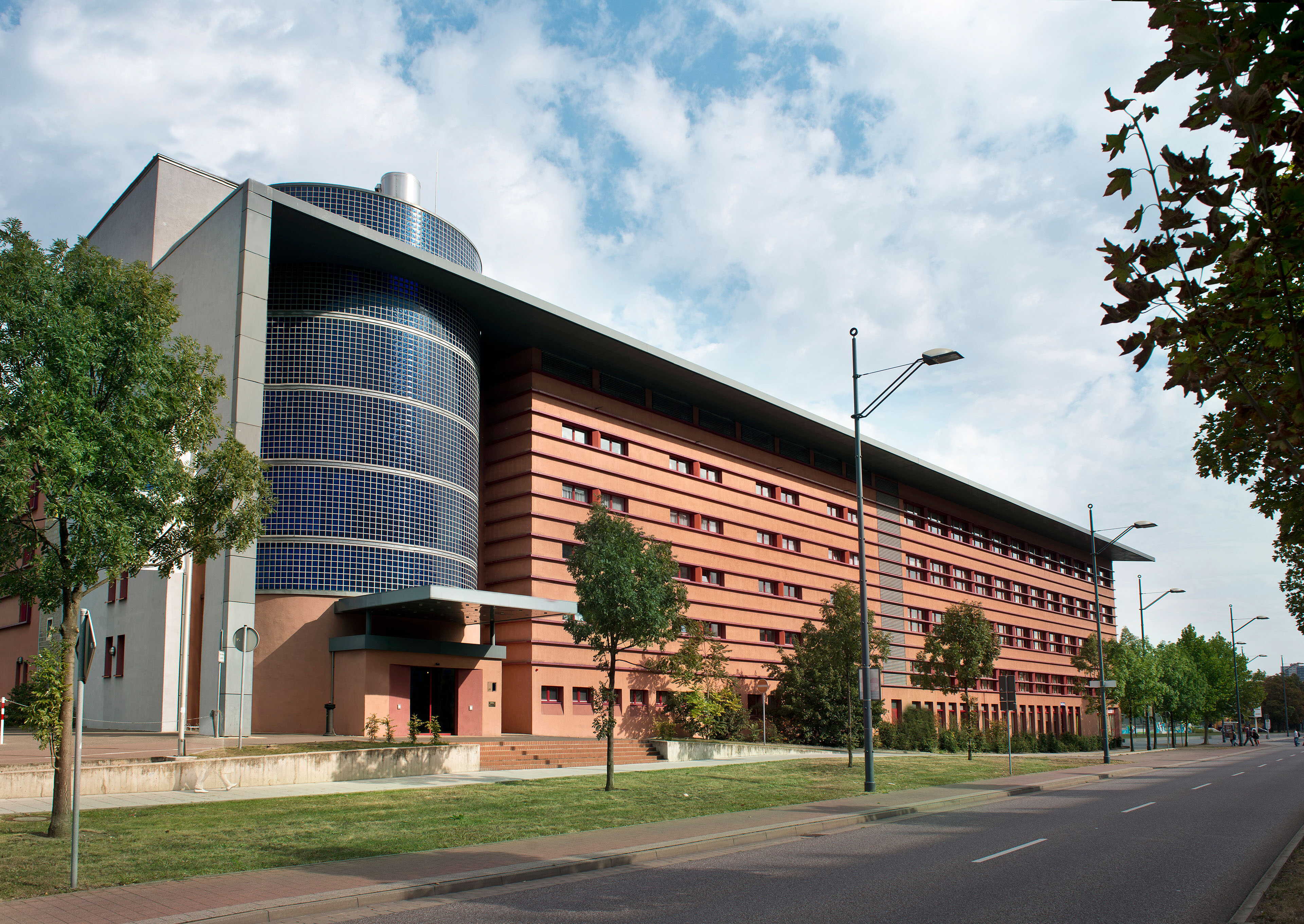 A modern building with a unique cylindrical glass section stands beside a tree-lined street on a cloudy day.