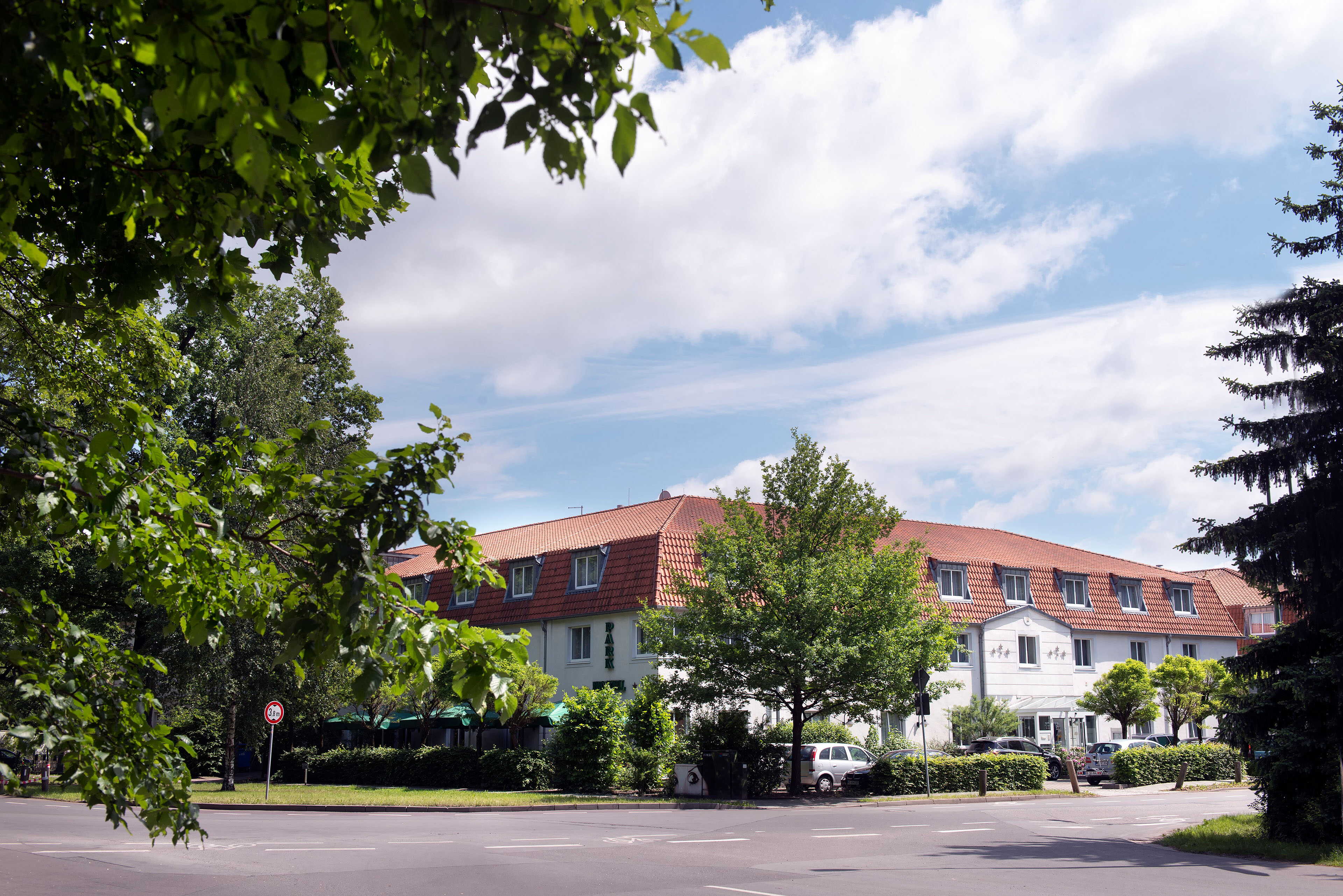 The image shows a large building with a red roof surrounded by lush trees and a clear blue sky.