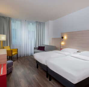 Helles Hotelzimmer mit zwei Betten, einem grauen Sofa und gelbem Stuhl bietet Komfort für Familienaufenthalte.