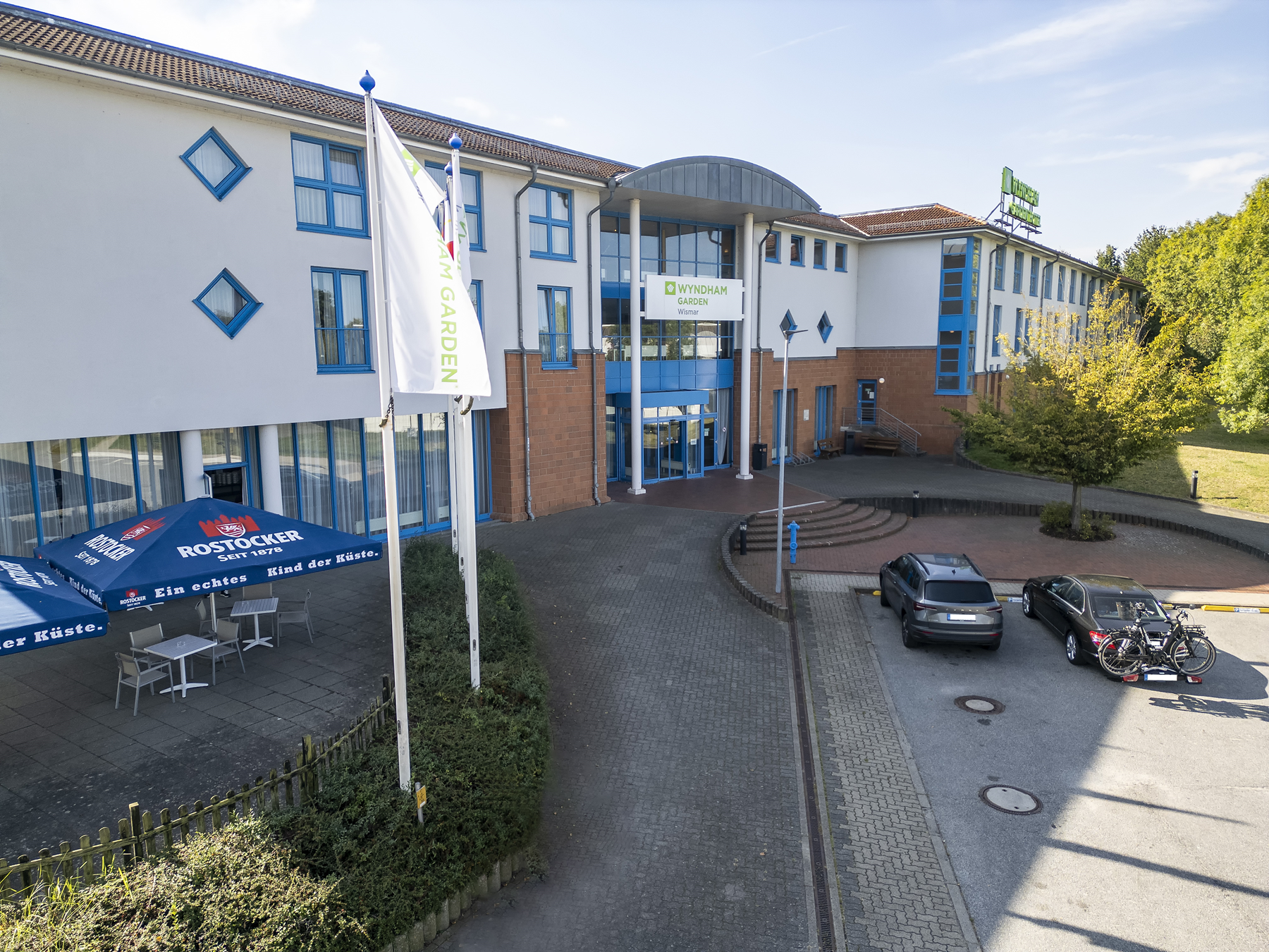 Aerial view of the Wyndham Garden hotel entrance with parked cars and patio seating under umbrellas.