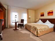 A cozy standard hotel room features a double bed, a small table with two chairs, and warm decor with scenic artwork.