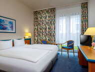 A cozy hotel room features a neatly made bed, vibrant floral curtains, and a small table with chairs by the window.