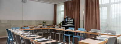 Ein moderner Konferenzraum ist für eine Tagung vorbereitet, mit U-förmig angeordneten Tischen und bereitgestelltem Schreibmaterial.