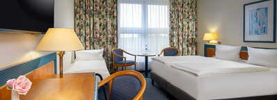 A cozy apartment room features a double bed, desk with a flower vase, and a small sitting area by a window with floral curtains.