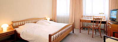 Ein gemütliches Doppelzimmer mit Holzmöbeln, Schreibtisch, Fernseher und großen Fenstern, die viel Licht hereinlassen.