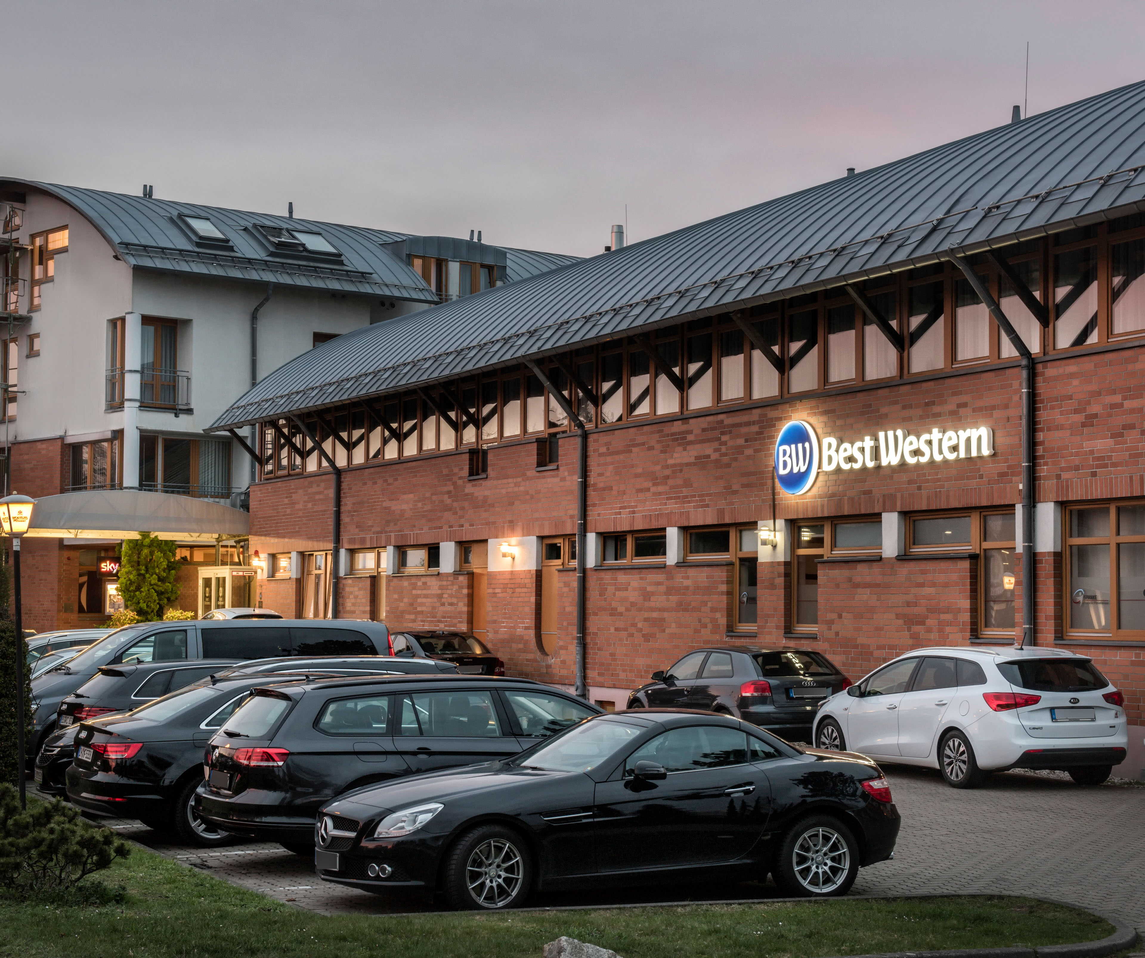 An evening shot of the Best Western Hotel Braunschweig Seminarius, showing the entrance area and car park of the hotel, with several cars parked on the tarmac area in front of the hotel and lanterns providing lighting.