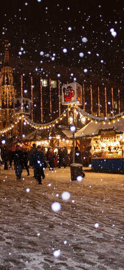 Pferdekutsche fährt durch den beleuchteten Weihnachtsmarkt im Schnee, während Menschen die festliche Atmosphäre genießen.