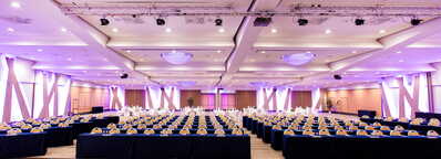A spacious conference room is set up with rows of tables and chairs, illuminated by purple lighting for a formal event.