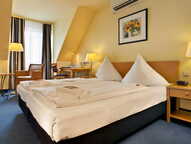 A cozy double room features a neatly made bed, warm lighting, and a small desk area, creating a welcoming atmosphere.