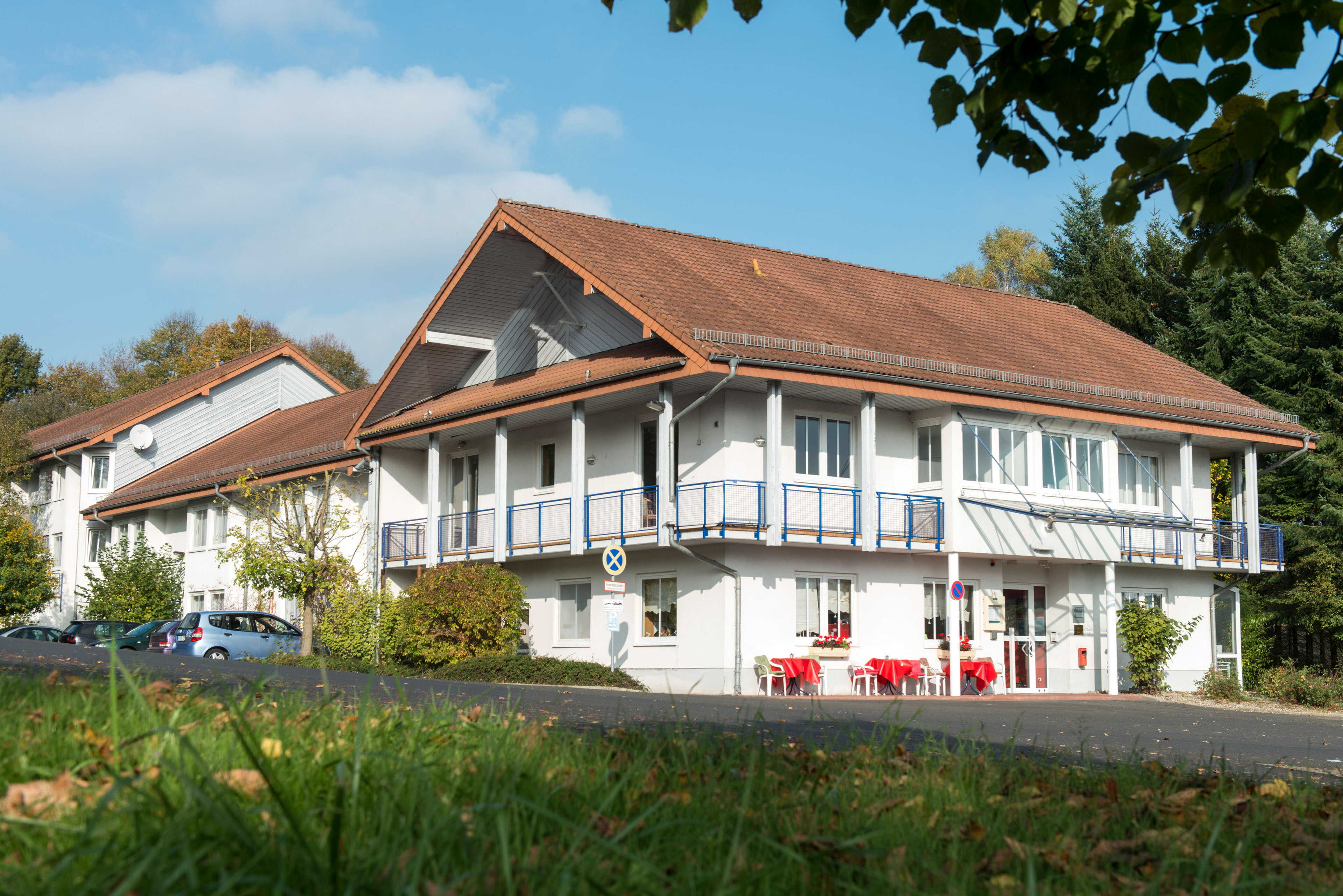 A two-story white building with a red-tiled roof stands amid trees and parked cars beneath a clear blue sky.