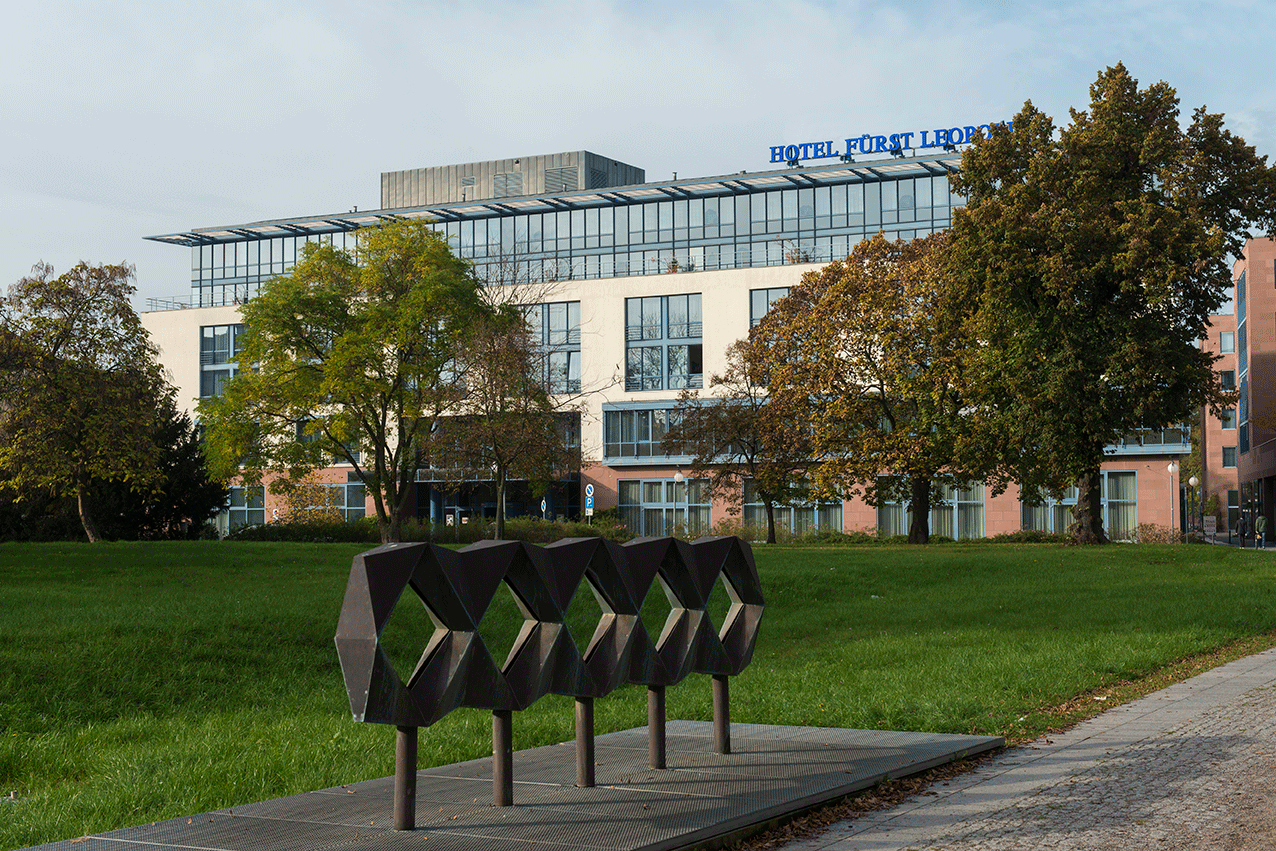 A modern hotel building with large windows stands behind a geometric sculpture and lush green lawn.