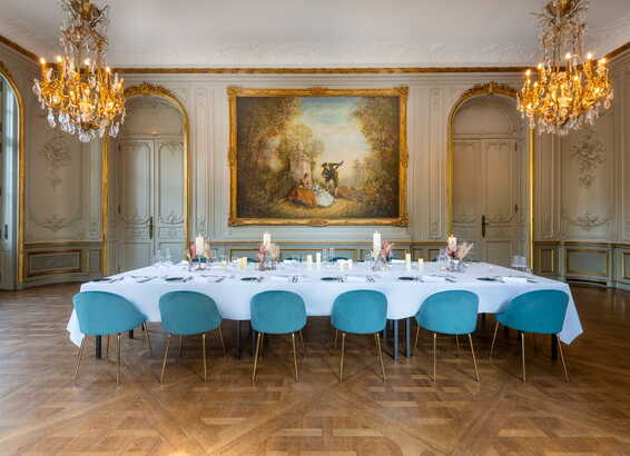 An elegant dining room features a long table with blue chairs, set under ornate chandeliers and a large classical painting.