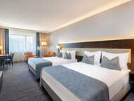 A modern hotel room features two neatly made beds with blue accents, a desk, and a window letting in natural light.