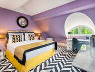 A stylish boutique room features a plush bed, purple walls, and a chevron-patterned floor under a round window.