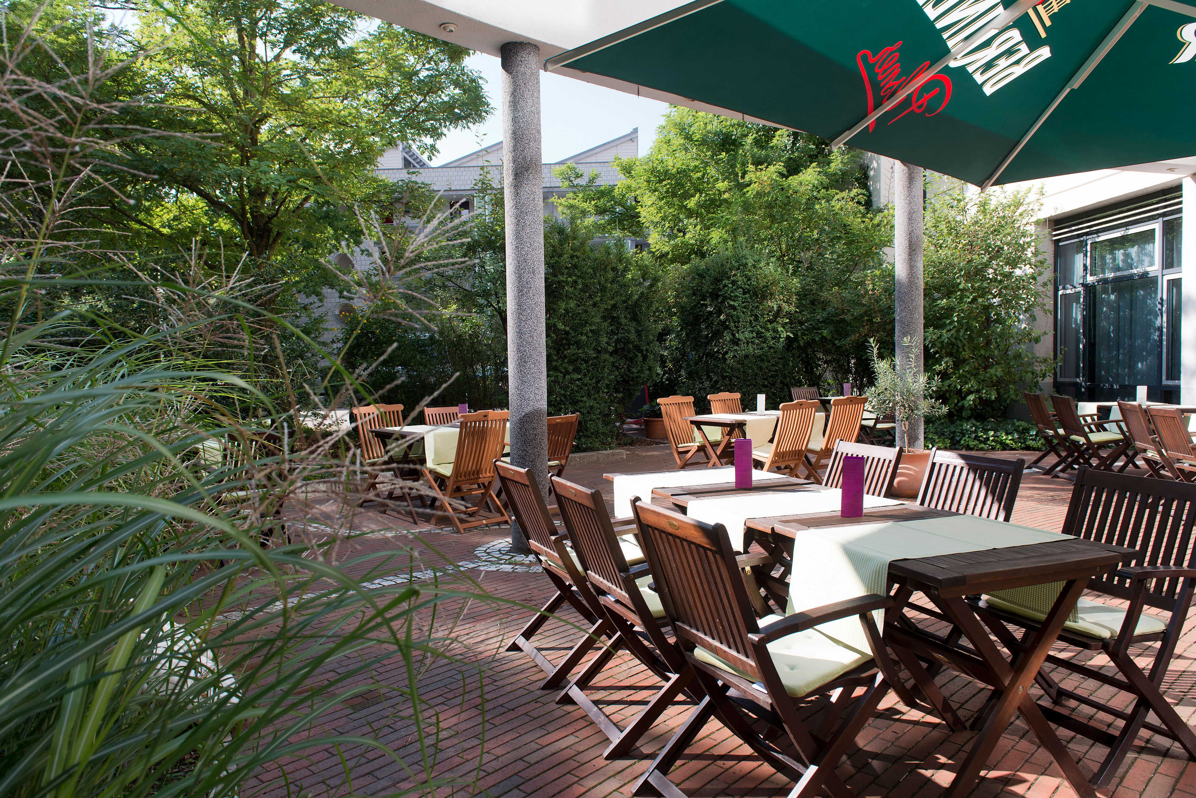 A garden terrace with wooden tables and chairs is shaded by green umbrellas, surrounded by lush greenery.