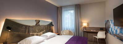 A stylish double room features a bed with a unique headboard, a purple throw, a desk with a chair, and a wall-mounted TV.