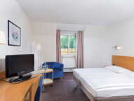 Helles Doppelzimmer mit einem gemütlichen Bett, einem Sessel, einem Schreibtisch mit Fernseher und einem Fenster mit Vorhang.