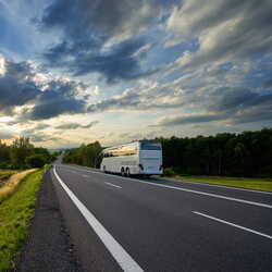 Ein weißer Reisebus fährt auf einer Landstraße in die Ferne, während die Sonne durch dramatische Wolken scheint.