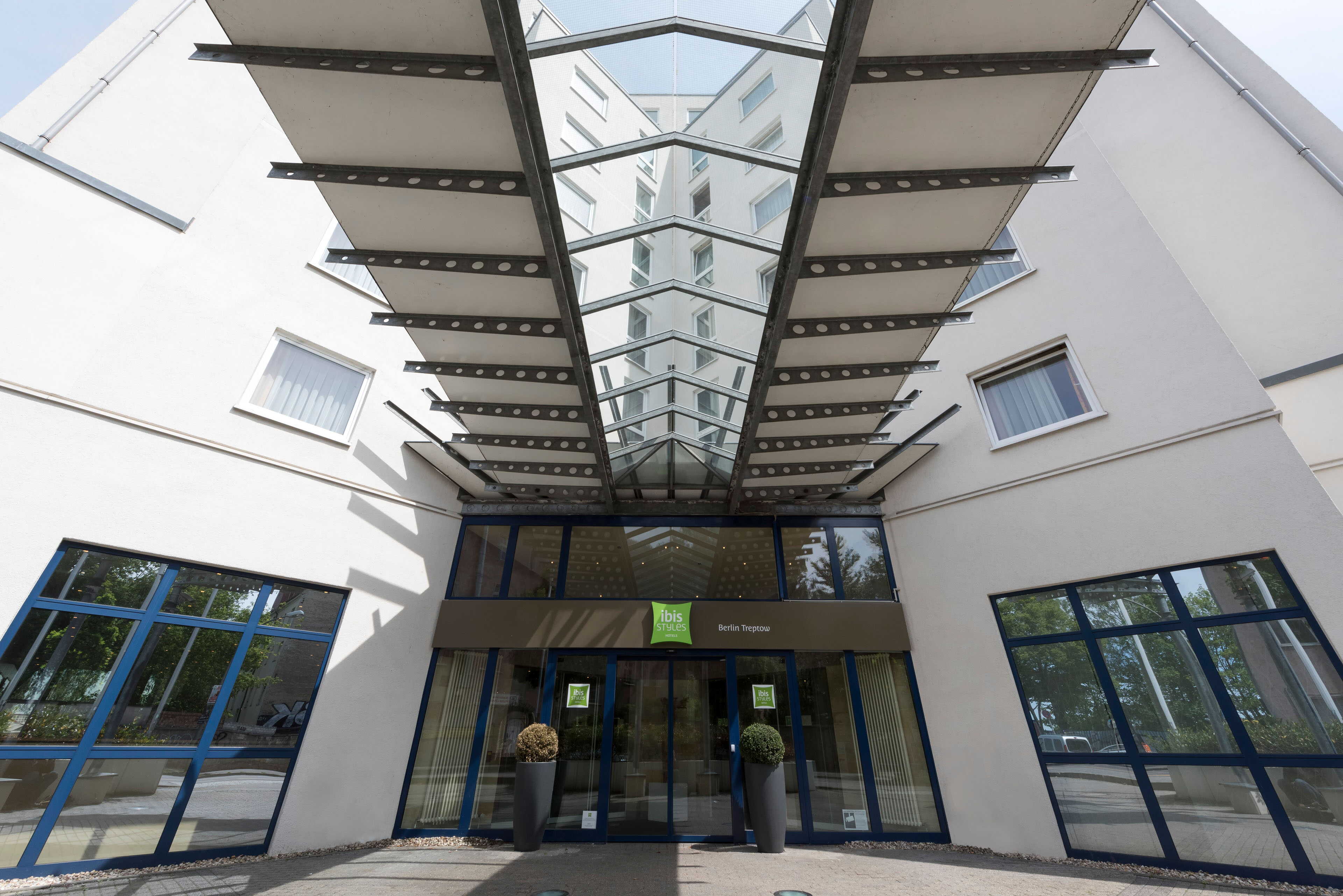 Modern hotel entrance with large glass canopy and blue-framed windows in Berlin Treptow, emphasizing sleek architecture.