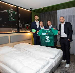 Vier Männer in Anzügen präsentieren ein grünes Handballtrikot in einem modern eingerichteten Hotelzimmer.