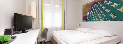 Ein modernes, helles Hotelzimmer mit Doppelbett, Flachbildfernseher und farbenfrohem Wandbild lädt zum Entspannen ein.