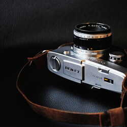 Eine klassische PEN-FT Kamera liegt mit einem braunen Lederriemen auf einem dunklen Hintergrund.