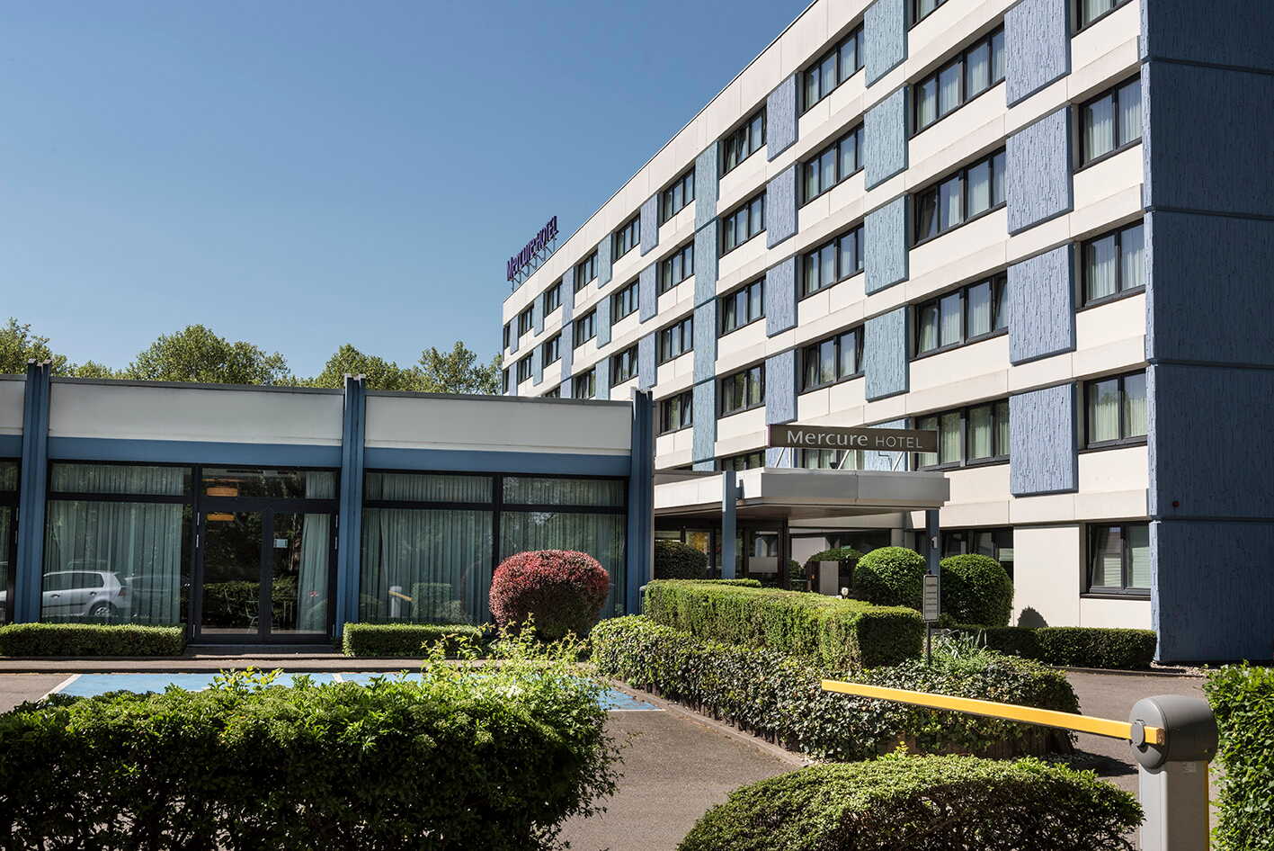 The exterior of a Mercure hotel features a modern facade and well-maintained hedges under a clear blue sky.