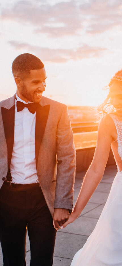 A couple in elegant attire joyfully holds hands on a rooftop with a stunning sunset city view.