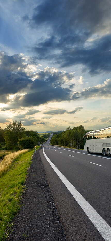 A white tour bus travels on a scenic highway at sunset, with dramatic clouds and lush greenery lining the road.