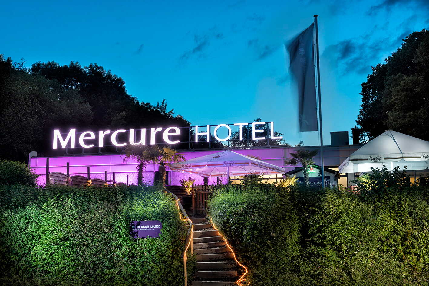 The exterior of a Mercure hotel is illuminated with vibrant lights against a twilight sky, featuring a welcoming entrance.