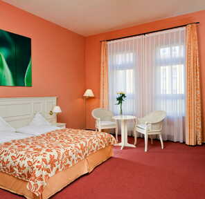 Gemütliches Zimmer mit einem großen Bett, einem kleinen Tisch mit zwei Stühlen und einem beruhigenden grünen Kunstwerk an der Wand.