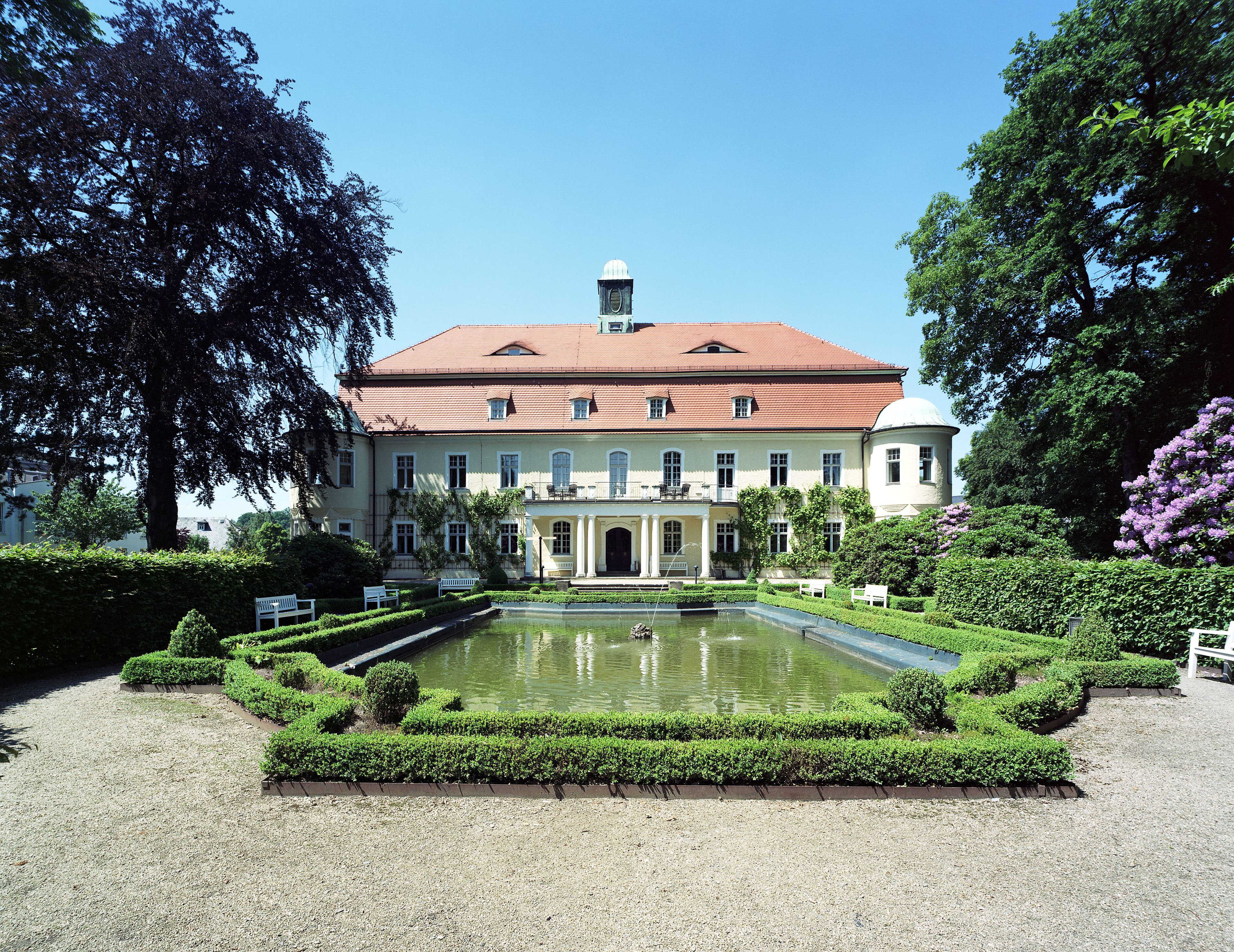 A grand baroque facade with a red roof overlooks a manicured garden and reflective pond under a clear blue sky.