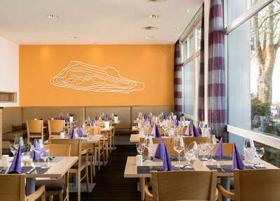 Ein stilvolles Restaurant mit gedeckten Tischen und violetten Servietten vor einem großen Fenster und einer orangen Wand.