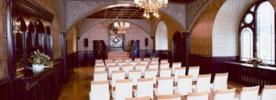 Weiße Stühle sind in einem prächtigen Rittersaal mit Kronleuchtern und dekorativen Wänden aufgereiht.