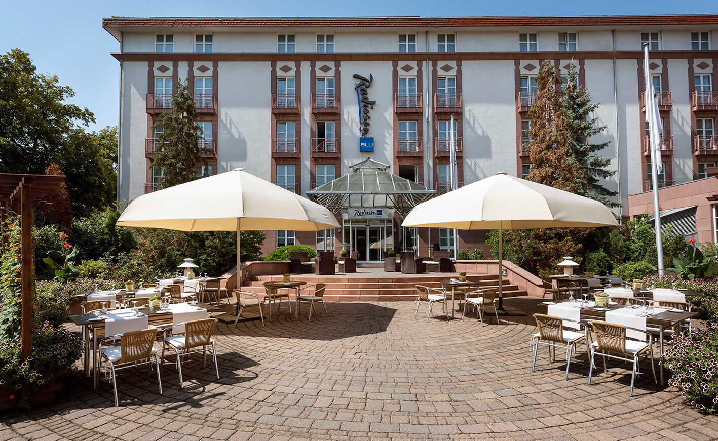 The exterior of a hotel features a charming patio with tables, chairs, and large umbrellas under a clear blue sky.