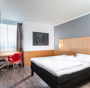 Ein modernes Einzelzimmer mit einem großen Bett, einem roten Stuhl am Schreibtisch und einem Kunstwerk an der Wand.