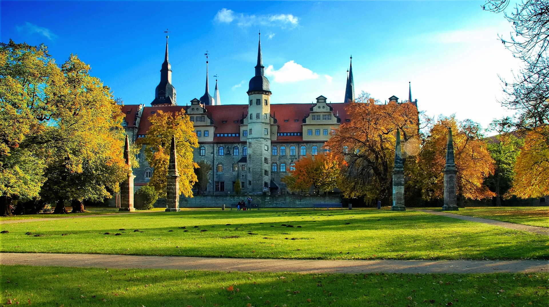 Das Schloss Merseburg strahlt im Sonnenlicht, umgeben von herbstlich gefärbten Bäumen und einer gepflegten Rasenfläche.
