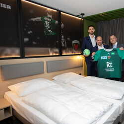 Vier Männer in Anzügen präsentieren ein grünes Handballtrikot in einem modern eingerichteten Hotelzimmer.